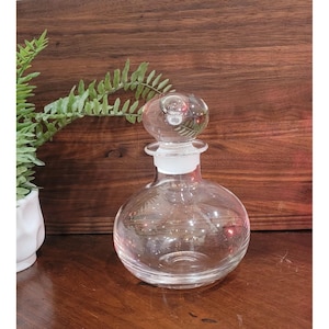May include: A clear glass perfume bottle with a round base and a clear stopper. The stopper has a round, bubble-like design. The bottle is on a wooden surface with a green plant in the background.