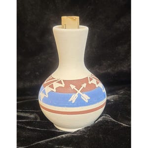 May include: A white ceramic vase with a narrow neck and a bulbous base. The vase is decorated with a band of blue, brown, and white geometric designs and crossed arrows. A cork stopper is visible at the top.