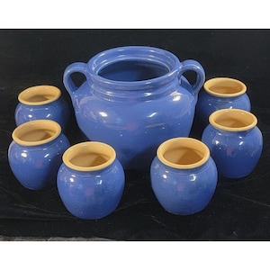 Könnte beinhalten: Ein Set aus blauer Keramik. Das Set besteht aus einem großen Topf mit Henkeln und sechs kleineren Töpfen. Die Innenseiten der Töpfe sind hellbeige, im Kontrast zum blauen Äußeren. Das Geschirr ist auf einer dunklen Oberfläche angeordnet.