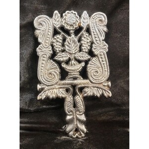 May include: Silver metal trivet with an ornate design of grapes, leaves, and flowers. The trivet has a decorative handle and is likely used to protect surfaces from hot cookware. The design is symmetrical and detailed, with a vintage aesthetic.