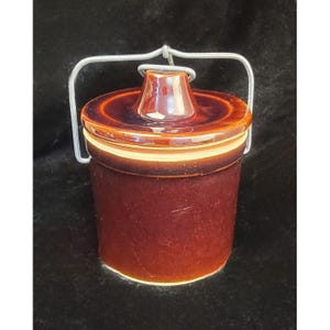 May include: A brown ceramic jar with a lid and a metal handle. The jar has a cylindrical shape with a slightly flared base. The lid is a darker brown and has a small knob on top. A white band separates the lid from the jar.