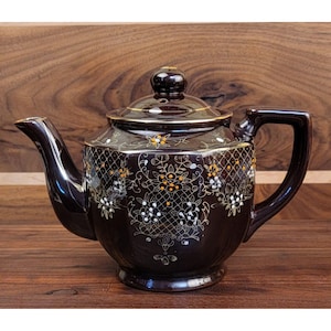 May include: A dark brown ceramic teapot with a lid and handle. The teapot is decorated with gold and white floral patterns and a lattice design. The spout and handle are a solid dark brown color.