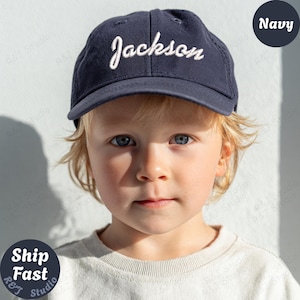 Puede incluir: Gorra de béisbol azul marino con el nombre "Jackson" bordado en escritura blanca. La gorra la lleva un niño pequeño con pelo rubio y ojos azules. La palabra "Navy" está en un círculo blanco en la esquina superior derecha.