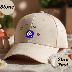 Peut inclure: Casquette de baseball beige avec une visière incurvée. La casquette présente des icônes colorées brodées et le mot "Stone" dans un cercle en haut à gauche. Les mots "Ship Fast" sont dans un cercle en bas à droite.