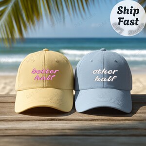May include: Two baseball caps, one yellow and one blue, are displayed. The yellow cap features the pink embroidered text "better half". The blue cap has the white embroidered text "other half". The background shows a beach scene with palm leaves.
