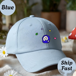 May include: A light blue baseball cap with embroidered designs, including a spiral, star, and heart. The word "Blue" is in a blue circle on the top left. The cap has a curved brim and a rounded crown.