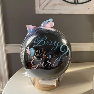 May include: A clear, spherical balloon filled with a black interior, featuring the text "Boy or Girl" in blue and pink script. The balloon is adorned with a light blue and pink ribbon bow at the top and rests on a circular, tan-colored base.