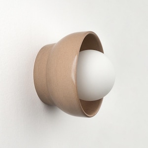 May include: A contemporary wall light with a beige ceramic shade and a spherical white bulb. The fixture is mounted on a white wall, demonstrating a minimalist design. The light is a simple, yet elegant addition to any interior.