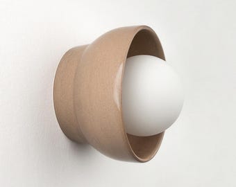May include: A contemporary wall light with a beige ceramic shade and a spherical white bulb. The fixture is mounted on a white wall, demonstrating a minimalist design. The light is a simple, yet elegant addition to any interior.