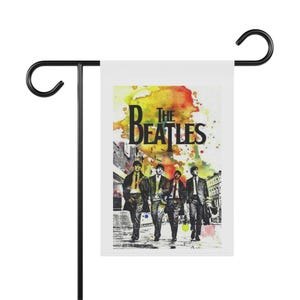 May include: A rectangular garden flag featuring a watercolor-style print of The Beatles walking on a street. The design includes vibrant yellow, orange, and green paint splatters above the band members, with the text THE BEATLES in black.