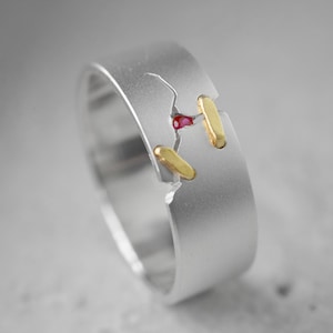 Puede incluir: Un anillo de plata con un diseño único. El anillo presenta una grieta con una pequeña gema roja y detalles dorados. El anillo tiene un acabado de metal cepillado y una estética moderna.