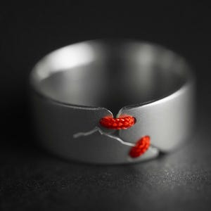 May include: A silver-colored ring with a unique design. The ring features two sections of bright red thread woven through the metal. The ring is a minimalist piece of jewelry.