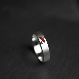 May include: A silver ring with a brushed finish. The ring has a cut-out design filled with red material. The ring is set against a dark background, highlighting its modern aesthetic.