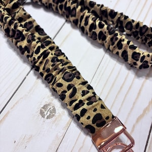 May include: A leopard print lanyard with a gold-colored metal clasp and key ring. The lanyard is made of a tan fabric with brown spots. The metal hardware is a shiny rose gold color.