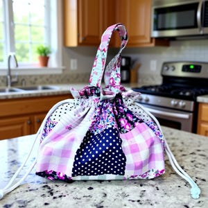 May include: A patchwork fabric lunch bag with a drawstring closure and a floral patterned handle. The bag features a mix of pink gingham, navy blue polka dots, and floral print fabrics. The bag is sitting on a granite countertop.