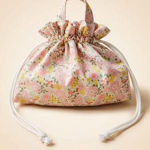 May include: A fabric drawstring pouch featuring a floral pattern with pink, yellow, and green blossoms on a light background. The bag has a gathered top, a single handle at the center, and two white cord drawstrings with knotted ends.