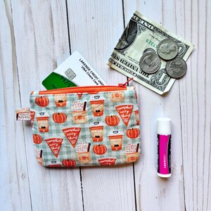May include: A small, patterned coin purse with an orange zipper, open to reveal a credit card. The purse features a repeating pattern of pumpkins, coffee cups, and the text "pumpkin everything". A dollar bill, coins, and lip balm are next to the purse.