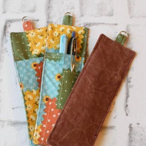 May include: Three fabric pen holders with a patchwork design. One is solid brown, and the others feature floral and geometric patterns in yellow, green, orange, and blue. Each has a metal ring and holds pens.