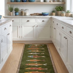 May include: A green rectangular rug with a repeating fish pattern in various colors, including orange, blue, and yellow. The rug is placed on a wooden floor in a kitchen setting, with white cabinets and shelves in the background.