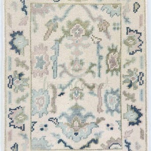May include: A rectangular rug with a cream-colored background and a detailed floral pattern in shades of blue, green, and mauve. The rug has a fringed edge and a central design framed by a border.