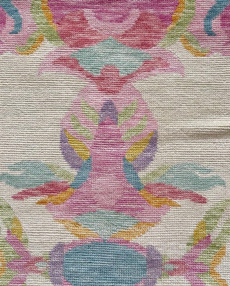 May include: A woven textile featuring a repeating symmetrical pattern in shades of pink, light blue, yellow, and green on an off-white background. The design consists of stylized floral and leaf motifs with a dense, textured, looped pile construction.