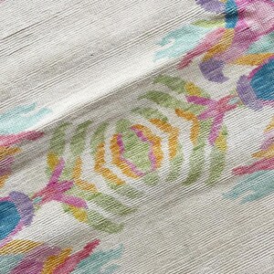 May include: A textile fabric featuring a textured off-white base with a repeating embroidered pattern in vibrant shades of pink, yellow, light blue, and green. The design consists of stylized floral or botanical motifs arranged in rows.
