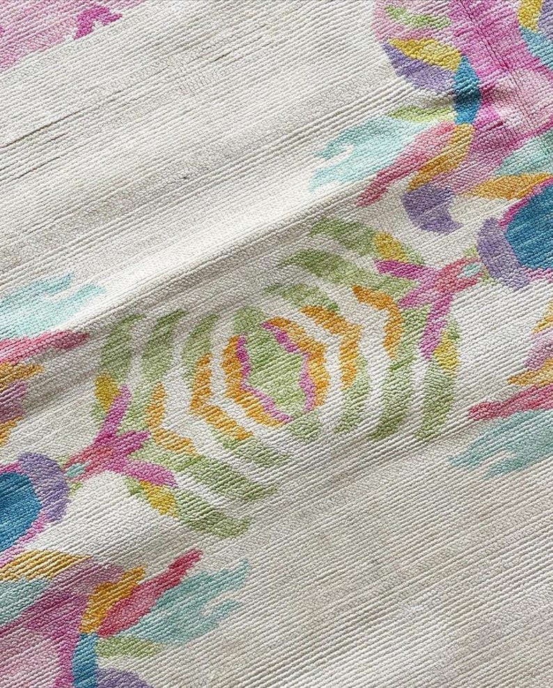 May include: A textile fabric featuring a textured off-white base with a repeating embroidered pattern in vibrant shades of pink, yellow, light blue, and green. The design consists of stylized floral or botanical motifs arranged in rows.
