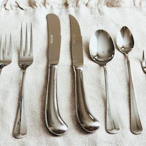 Pode incluir: Um conjunto de talheres com acabamento prateado, incluindo um garfo, uma faca e uma colher. Os talheres estão dispostos sobre uma toalha de mesa de linho bege.