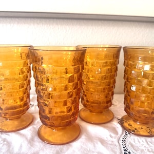 May include: A set of four amber-colored glass tumblers featuring a textured, block-like geometric pattern on the exterior. Each glass has a cylindrical body that tapers slightly toward a circular pedestal base, creating a uniform appearance.