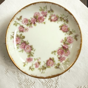 May include: A round porcelain saucer featuring a white base with a delicate floral pattern of pink flowers and green leaves arranged in a circular wreath. The rim of the saucer is finished with a worn gold-toned metallic trim.