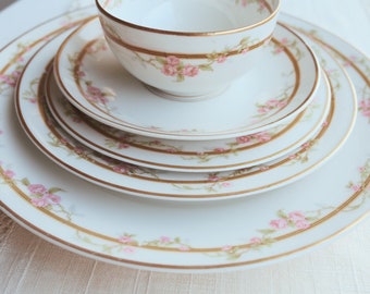 May include: A set of four white porcelain plates with a delicate pink floral pattern and gold trim. The plates are stacked on top of each other, with a matching teacup and saucer on top.