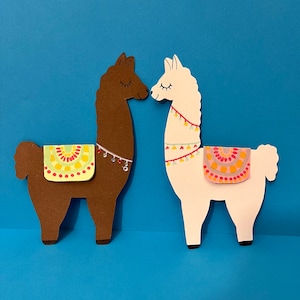 K&ouml;nnte beinhalten: Zwei Papier-Ausschnitte von Lamas, die sich gegen&uuml;berstehen. Das Lama links ist braun mit einem gr&uuml;n-gelb-roten Sattel. Das Lama rechts ist wei&szlig; mit einem rosa-gelb-orangenen Sattel.