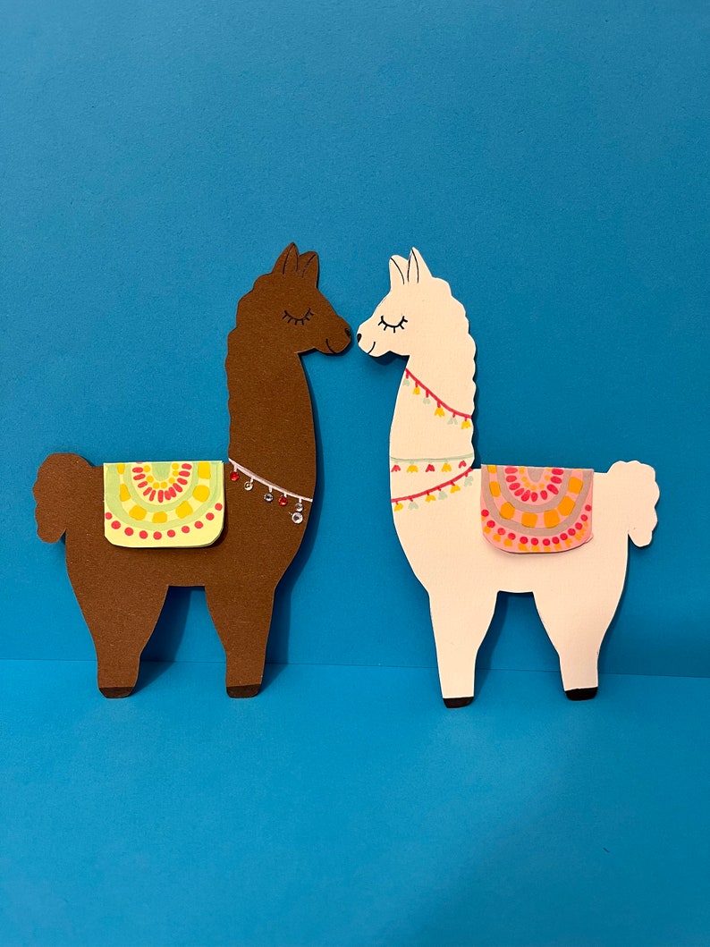 K&ouml;nnte beinhalten: Zwei Papier-Ausschnitte von Lamas, die sich gegen&uuml;berstehen. Das Lama links ist braun mit einem gr&uuml;n-gelb-roten Sattel. Das Lama rechts ist wei&szlig; mit einem rosa-gelb-orangenen Sattel.