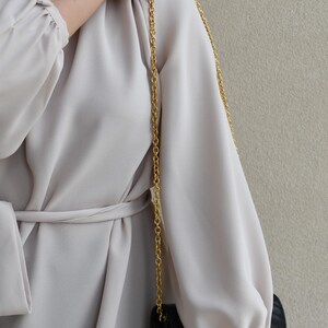 A person wearing a white dress with a gold chain strap, holding a black purse, and standing against a beige wall.