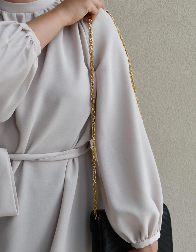 A person wearing a white dress with a gold chain strap, holding a black purse, and standing against a beige wall.