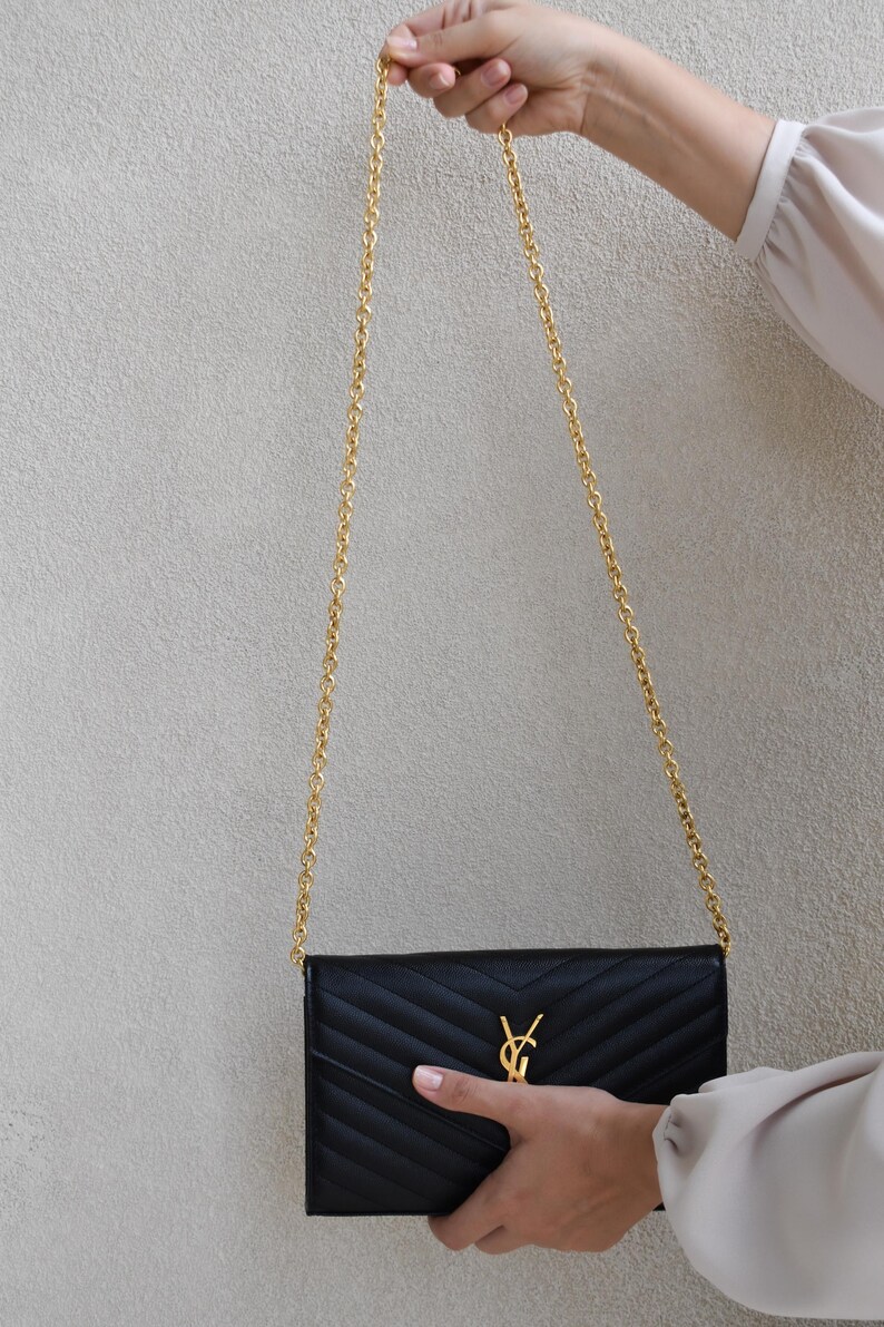 A person's hand holding a black Yves Saint Laurent wallet with a gold chain strap.