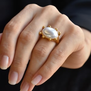 May include: A gold ring featuring a large, oval, white pearl. The ring has a gold band with small, decorative gold accents around the pearl. The ring is on a hand with manicured nails, set against a dark background.