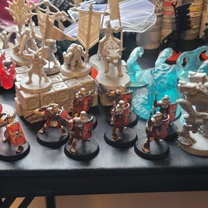 May include: A collection of miniature figurines, including soldiers with shields, fantasy creatures, and figures with banners. The miniatures are in various colors, including red, blue, and gray, and are arranged on a dark surface.