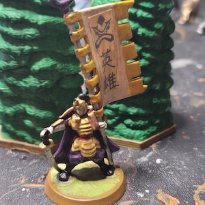 May include: A miniature figure of a warrior holding a banner with Japanese characters. The banner is yellow and brown with gold trim. The figure is wearing purple and gold armor and is holding a sword.