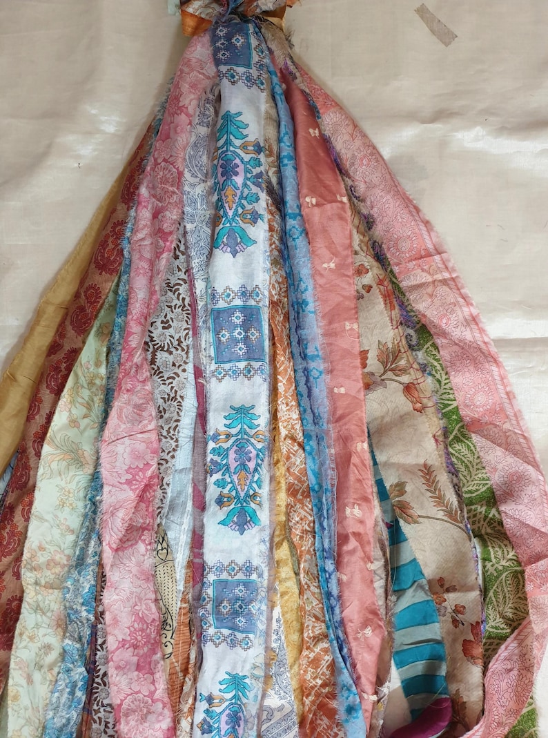 May include: A bundle of colorful fabric strips with various patterns, including floral, geometric, and paisley designs. The strips are tied together at the top and hang down in a cascade.