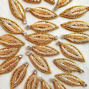 May include: Close-up of multiple gold-colored leaf-shaped embellishments. Each leaf has a detailed, textured design with a small, clear crystal at the tip. The leaves are arranged on a white surface.