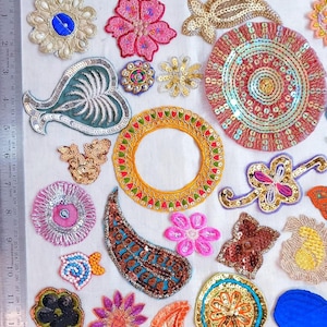 May include: A collection of assorted embroidered and sequined fabric patches in various shapes and colors, including floral, paisley, and circular designs, arranged on a white surface next to two metal rulers showing measurements in inches and centimeters.