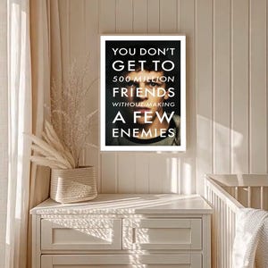 May include: A framed poster with the text "YOU DON'T GET TO 500 MILLION FRIENDS WITHOUT MAKING A FEW ENEMIES" in white against a black background. The poster features a person's face. The poster is hanging on a white wall.