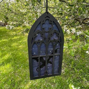 May include: Black gothic-style window hanging decoration with a dragon silhouette inside. The window has a dark blue background with white star-like accents. The decoration is hanging from a black string in front of a green grassy background.