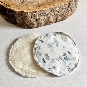 May include: Two reusable facial rounds. One is a soft, cream-coloured terry cloth. The other is white with a floral and dragonfly pattern in blue and green. The rounds are circular and sit on a white surface.