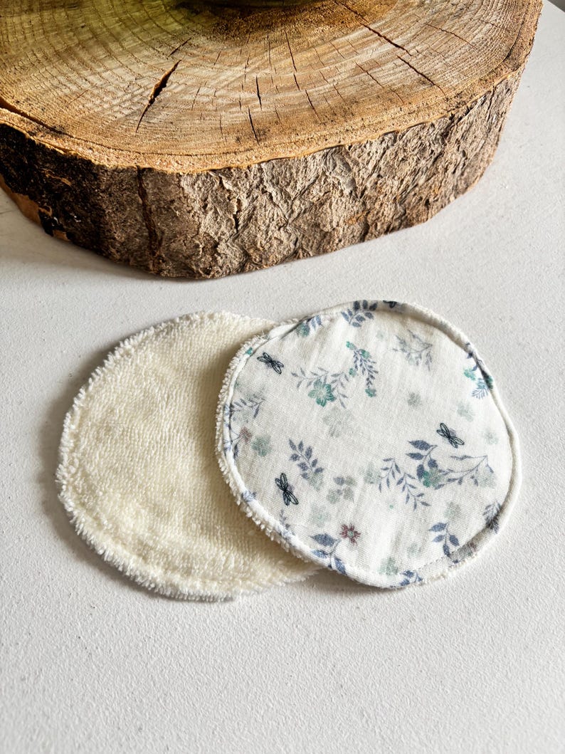 May include: Two reusable facial rounds. One is a soft, cream-coloured terry cloth. The other is white with a floral and dragonfly pattern in blue and green. The rounds are circular and sit on a white surface.