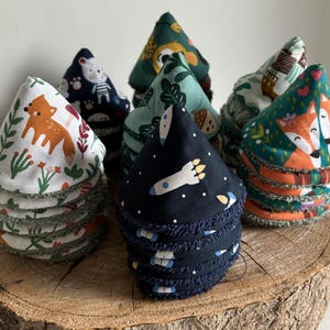 Peut inclure: Une collection de cotons démaquillants réutilisables faits à la main, avec divers motifs colorés. Les motifs incluent des renards, des fusées et des créatures de la forêt. Les cotons sont empilés sur une surface en bois, mettant en valeur leurs imprimés et textures.