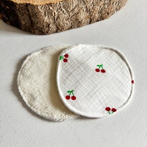 May include: Two circular reusable facial cleansing pads. One is white terry cloth, the other is white with a cherry print. The cherries are red with green stems. The pads are on a white surface.
