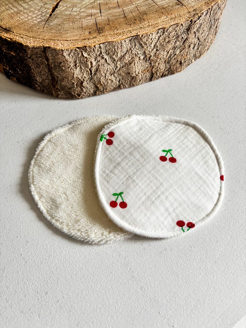 May include: Two circular reusable facial cleansing pads. One is white terry cloth, the other is white with a cherry print. The cherries are red with green stems. The pads are on a white surface.
