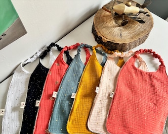 May include: A selection of baby bibs in shades of white, black, coral, blue, yellow, and pink. Each bib is made from a soft, textured material and decorated with small, gold star-shaped details. The bibs have a rounded shape and a neckband.